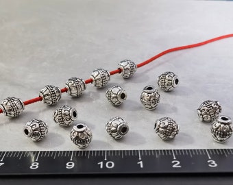 May include: A collection of silver-toned metal beads with intricate embossed patterns, featuring a central hole. Five beads are threaded onto a thin red cord, while others are scattered nearby. A black ruler shows measurements in centimeters.