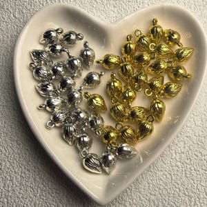Könnte beinhalten: Eine herzförmige Schale mit silbernen und goldfarbenen Metall-Eichel-Anhängern. Die Anhänger haben eine strukturierte Oberfläche und eine kleine Schlaufe zum Befestigen an Schmuck oder anderen Bastelarbeiten. Die Anhänger sind in zwei verschiedenen Gruppen angeordnet.