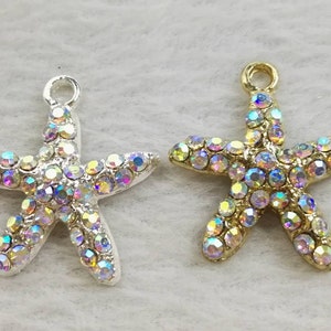 May include: Two starfish charms, one in a silver-tone metal and one in a gold-tone metal, each featuring a top loop for hanging and a surface densely encrusted with small, iridescent, multi-colored rhinestones that reflect light in various hues.