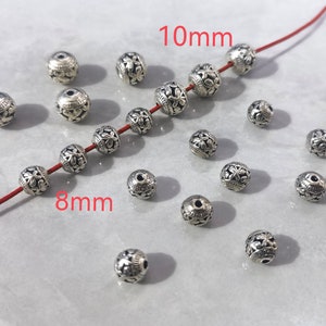 May include: A collection of round silver-toned metal beads featuring an intricate floral relief pattern. Some beads are strung on a thin red cord, while others are loose. The image includes text labels indicating sizes of 8mm and 10mm.