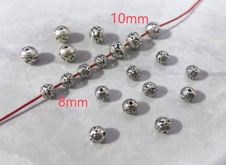May include: A collection of round silver-toned metal beads featuring an intricate floral relief pattern. Some beads are strung on a thin red cord, while others are loose. The image includes text labels indicating sizes of 8mm and 10mm.