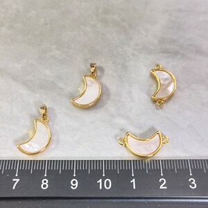 May include: Four gold-toned crescent moon pendants with a white, pearlescent inlay. The pendants vary in design, with some featuring a single loop for hanging and others with loops on both ends for connecting. The moons are set against a gray surface.