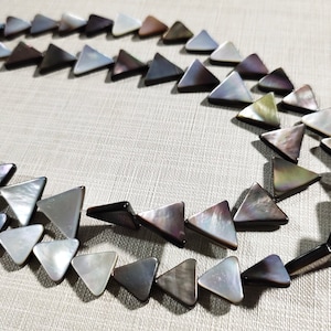 Puede incluir: Collar con cuentas triangulares iridiscentes en tonos grises, negros y blancos. Las cuentas están dispuestas en un patrón curvo, creando un diseño único y llamativo. El collar probablemente está hecho de materiales naturales.