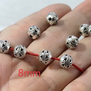 May include: A collection of round, silver-toned metal beads featuring intricate filigree patterns and a central beaded band. Four beads are threaded onto a red cord, while three others are loose. The text 8mm is visible in red.