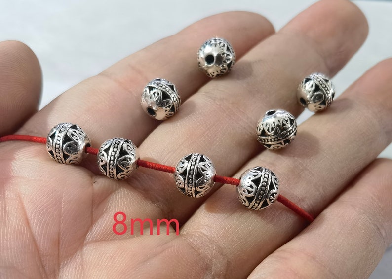 May include: A collection of round, silver-toned metal beads featuring intricate filigree patterns and a central beaded band. Four beads are threaded onto a red cord, while three others are loose. The text 8mm is visible in red.