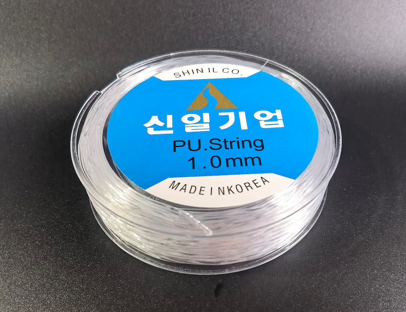 May include: A spool of clear 1.0mm PU string, made in Korea. The label reads "Shin Il Co.  PU.String 1.0mm Made in Korea" in Korean and English.