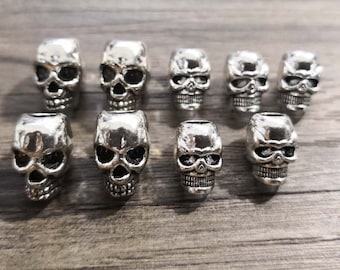 Puede incluir: Un conjunto de nueve cuentas de metal en tono plateado con forma de calavera, dispuestas en dos filas. Cada cuenta presenta un diseño tridimensional detallado de una calavera humana con cuencas oculares huecas, nariz y dientes definidos.