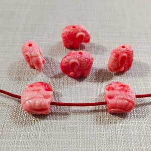 May include: Six pink carved elephant beads with a hole through the top. The beads are strung on a red cord.