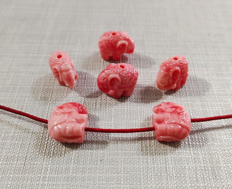 May include: Six pink carved elephant beads with a hole through the top. The beads are strung on a red cord.