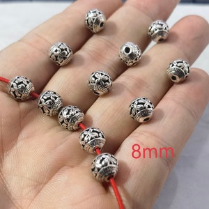 May include: A collection of spherical silver-toned metal beads, each measuring 8mm in diameter. The beads feature a detailed floral relief pattern and a central hole for threading. Several beads are strung on a thin red cord, while others are loose.
