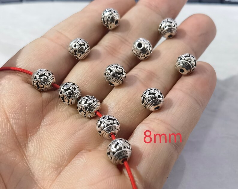 May include: A collection of spherical silver-toned metal beads, each measuring 8mm in diameter. The beads feature a detailed floral relief pattern and a central hole for threading. Several beads are strung on a thin red cord, while others are loose.