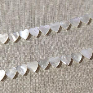 May include: Two strands of white heart-shaped shell beads on a beige fabric background.