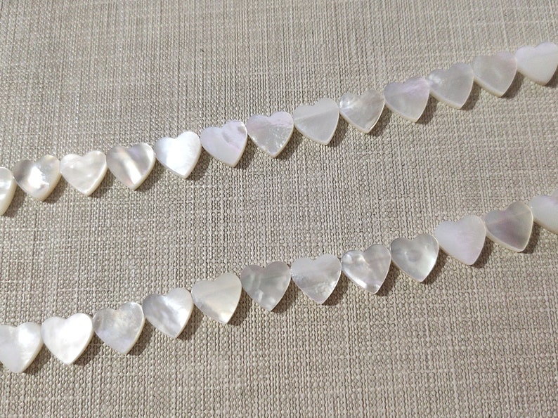 May include: Two strands of white heart-shaped shell beads on a beige fabric background.