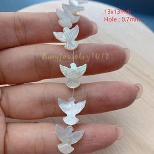 May include: A string of white mother-of-pearl beads carved into bird shapes. Each bead measures 13x13mm with a 0.7mm hole. The iridescent, flat-surfaced bird charms are arranged vertically on a thin thread, showcasing a natural pearlescent finish.