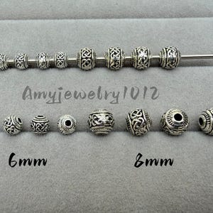 Könnte beinhalten: Eine Sammlung von silberfarbenen Metallperlen mit komplizierten keltischen Knotenmustern. Die Perlen sind in zwei Reihen angeordnet, einige auf einem Metallstab aufgereiht. Der Text Amyjewelry1012 sowie die Größenangaben 6mm und 8mm sind sichtbar.