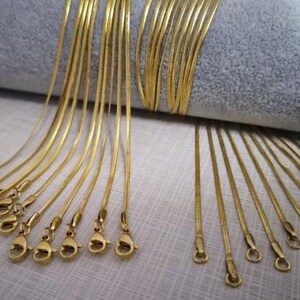 May include: A collection of gold snake chain necklaces with clasps. The chains are arranged in a fan shape on a gray fabric background.