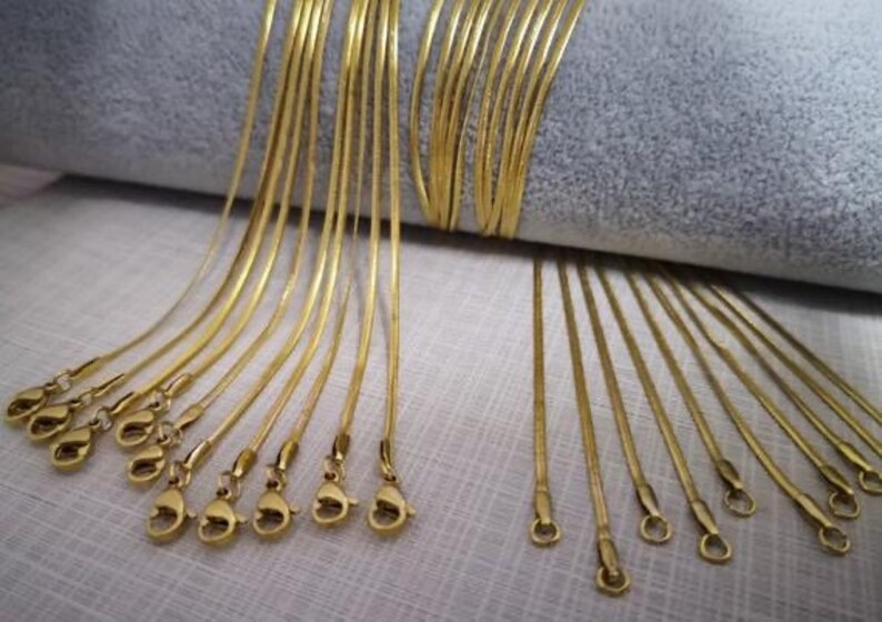 May include: A collection of gold snake chain necklaces with clasps. The chains are arranged in a fan shape on a gray fabric background.