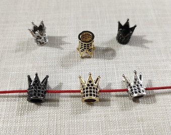 May include: A collection of six crown-shaped beads in silver, gold, and black metal finishes. Each bead features a hollow center and is encrusted with small, dark, sparkling stones. Three of the beads are threaded onto a thin, horizontal red cord.