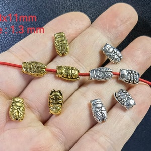 Op de afbeelding: Kleine metalen kralen in de vorm van cicaden in goud- en zilverkleuren. De kralen meten 6,8x11 mm met een gat van 1,3 mm. Sommige kralen zijn geregen aan een dun rood koord, terwijl andere los liggen en gedetailleerde insectentexturen tonen.