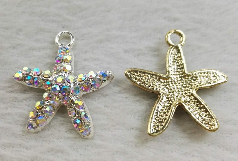 May include: Two starfish charms with a loop at the top. The left charm is silver-toned and covered in iridescent, multi-colored rhinestones. The right charm is gold-toned with a textured, metallic finish on the front surface of the starfish shape.