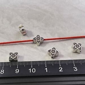 May include: Five silver metal beads with a decorative floral design. The beads are strung on a red cord and are displayed on a ruler with markings from 8 to 10 inches.