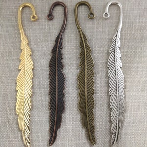 May include: Four metal feather-shaped bookmarks arranged vertically. Each bookmark features a curved hook at the top with a small circular loop. The set includes one shiny gold, one dark copper, one antique bronze, and one silver-toned finish.