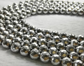 May include: A strand of silver-toned faceted beads, arranged in a circle on a white surface. The beads are round and have a metallic sheen.