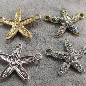 Op de afbeelding: Vier zeestervormige bedels met twee zijdelingse ringen voor bevestiging. Twee bedels hebben een getextureerde metallic afwerking in goud en zilver, terwijl de andere twee zijn ingelegd met iriserende, veelkleurige strass-steentjes.