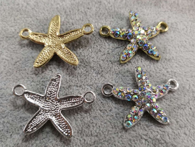 Op de afbeelding: Vier zeestervormige bedels met twee zijdelingse ringen voor bevestiging. Twee bedels hebben een getextureerde metallic afwerking in goud en zilver, terwijl de andere twee zijn ingelegd met iriserende, veelkleurige strass-steentjes.