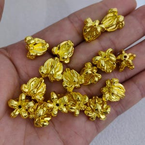 May include: A collection of small, gold-colored flower-shaped beads. The beads vary in size and design, with some featuring multiple petals and others appearing more compact. The beads are displayed against a neutral background.