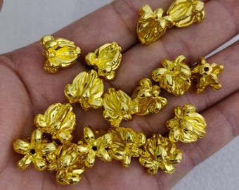 May include: A collection of small, gold-colored flower-shaped beads. The beads vary in size and design, with some featuring multiple petals and others appearing more compact. The beads are displayed against a neutral background.