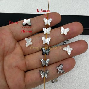 May include: A collection of butterfly-shaped beads in white and dark gray, featuring a 10mm width, 8mm height, and 0.8mm thickness as indicated by red text. The beads are arranged on a thin gold wire, with some loose beads visible.