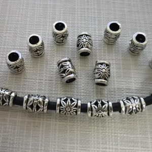 K&ouml;nnte beinhalten: Silberne Metallperlen mit floralem Design. Die Perlen sind auf einer schwarzen Schnur aufgef&auml;delt.