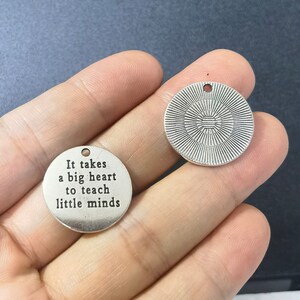 Puede incluir: Dos dijes de metal plateado. Un dije es redondo y tiene el texto "Se necesita un gran coraz&oacute;n para ense&ntilde;ar a las mentes peque&ntilde;as". El otro dije es redondo y tiene un dise&ntilde;o decorativo.