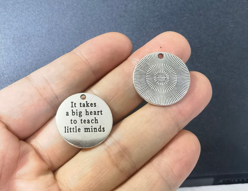 Puede incluir: Dos dijes de metal plateado. Un dije es redondo y tiene el texto "Se necesita un gran coraz&oacute;n para ense&ntilde;ar a las mentes peque&ntilde;as". El otro dije es redondo y tiene un dise&ntilde;o decorativo.