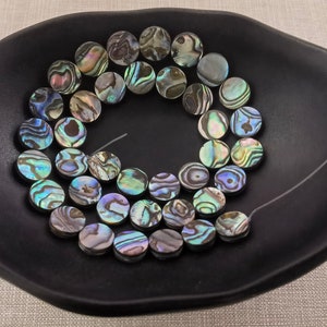 May include: A selection of iridescent abalone shell beads, each approximately 2.5 cm in diameter, presented in a black ceramic dish. The beads are strung on a thin, clear thread, highlighting their natural, swirling patterns of blues, greens, and browns.