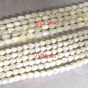 May include: Two strands of off-white, iridescent shell beads carved into tulip shapes. The top strand features beads labeled 8x11mm, while the bottom strand features smaller beads labeled 7x8mm. The text Amyjewelryvn is visible in the center.