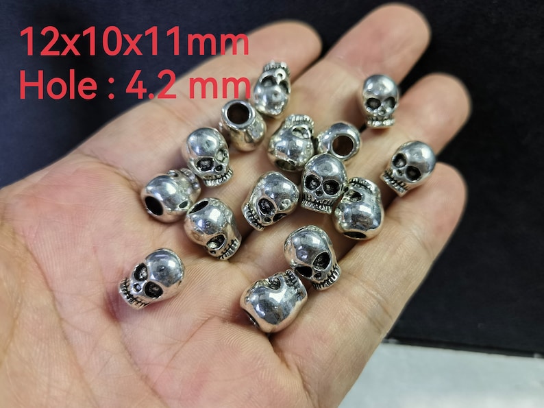 May include: A collection of silver-toned metal skull beads, each measuring 12x10x11mm with a 4.2mm hole. The beads feature detailed eye sockets, nasal cavities, and teeth, with a polished finish that reflects light across their rounded surfaces.