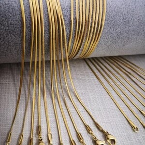 May include: A group of 20 gold snake chain necklaces with lobster claw clasps. The chains are laid out in a fan shape on a light gray surface.
