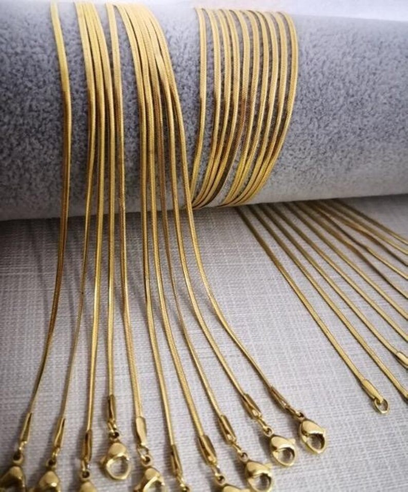 May include: A group of 20 gold snake chain necklaces with lobster claw clasps. The chains are laid out in a fan shape on a light gray surface.