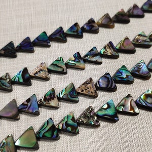 Puede incluir: Una hebra de cuentas triangulares de concha de abulón iridiscente. Las cuentas son una mezcla de colores verde, azul y marrón.