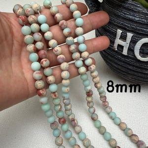 May include: Strands of 8mm round beads in shades of pale blue, beige, and red. The beads have a matte finish and are strung together. The image shows multiple strands of the beads, with some held in a hand.
