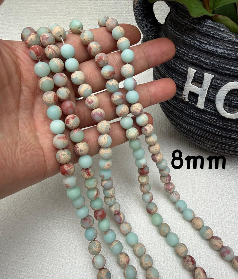 May include: Strands of 8mm round beads in shades of pale blue, beige, and red. The beads have a matte finish and are strung together. The image shows multiple strands of the beads, with some held in a hand.