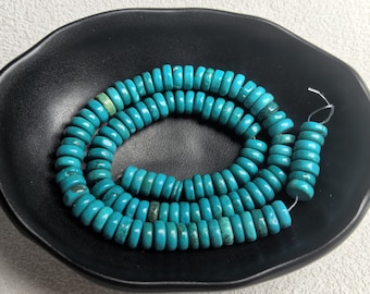 May include: A strand of turquoise-colored, disc-shaped beads. The beads are strung together, forming a necklace or bracelet. The beads are a vibrant blue-green color and are displayed on a black, shallow dish.