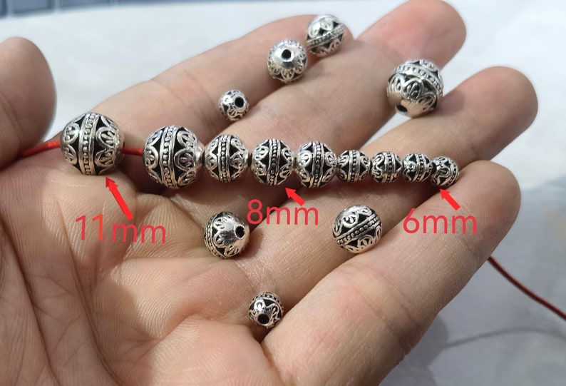May include: A collection of round, silver-toned metal beads featuring intricate filigree patterns and black recessed detailing. The beads are shown in various sizes, with red text labels indicating specific diameters of 11mm, 8mm, and 6mm.