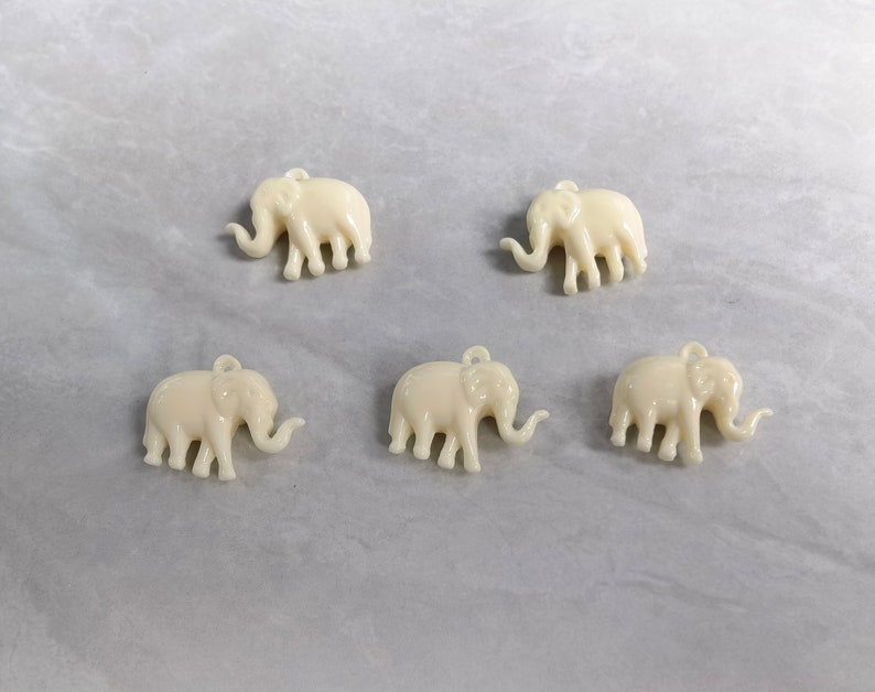May include: Five white plastic elephant charms with a loop for hanging. The elephants are all facing the same direction and have a raised trunk.