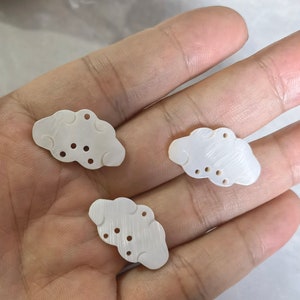 Op de afbeelding: Drie witte schelpvormige wolkvormige bedels met uitgesneden details. De bedels hebben een gladde, gepolijste afwerking.