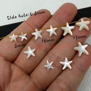 Könnte beinhalten: Eine Sammlung weißer sternförmiger Perlen mit glatter, perlmuttartiger Oberfläche. Die Perlen sind in verschiedenen Größen von 8mm, 10mm und 12mm dargestellt. Ein dünner goldener Draht führt durch die Perlen, Text weist auf ein 0,8mm Seitenloch hin.