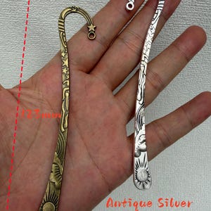 May include: Two metal bookmarks, one in antique bronze and one in antique silver, each featuring a curved top with a star and a sun motif at the base. The bookmarks measure 123 mm in length and 23 mm in width, with celestial patterns along the stems.