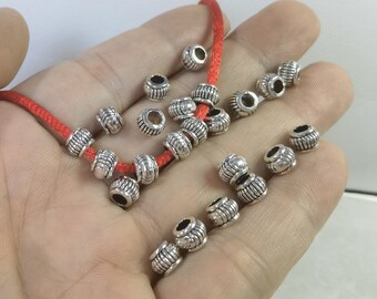 Può includere: Una collezione di piccole perline metalliche rotonde color argento, caratterizzate da una superficie scanalata e testurizzata con un foro centrale. Diverse perline sono infilate su un cordino rosso vivo, mentre altre sono sparse vicino.